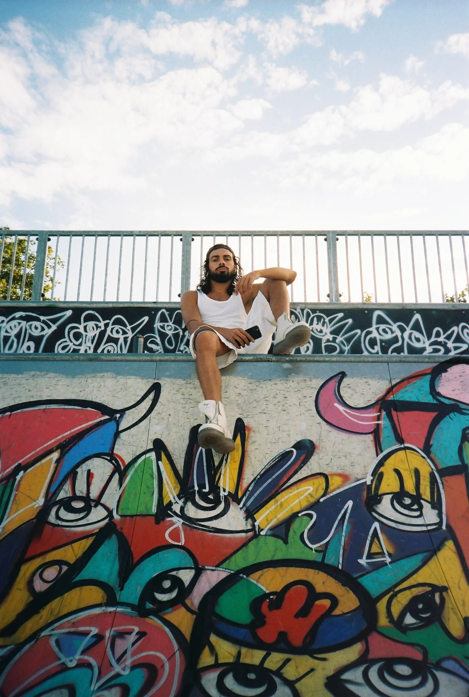 Portrait de P4NEL, assis en haut d'une rampe de skatepark couverte de graffitis colorés, sous un ciel bleu